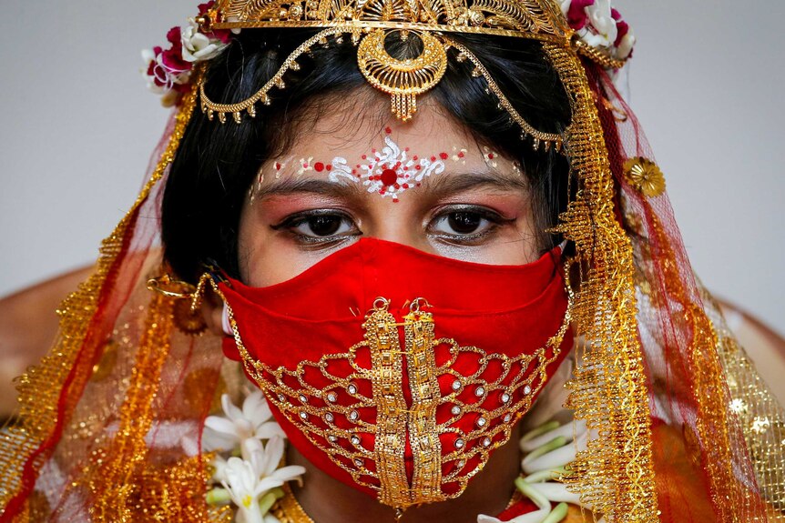 An Indian girl in a red face mask with gold headdress, veil and flowers in her hair