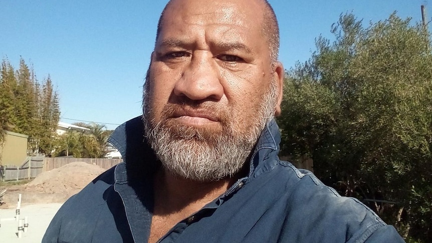 Head and shoulders selfie image of a man in a blue shirt on a building site.