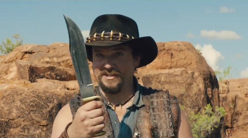 A screenshot shows a Crocodile Dundee-inspired character brandishing a knife in an outback scene.