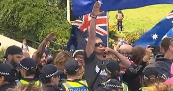 Surrounded by men wearing black and police officers, one man stands and raises arm in Nazi salute.