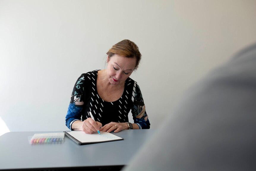 A woman in a plain room writes in a notebook on a table.