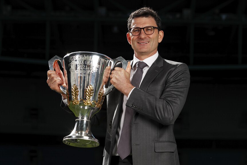 A man in a suit holds the AFL premiership cup aloft.