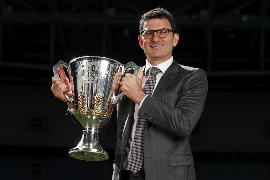 A man in a suit holds the AFL premiership cup aloft.