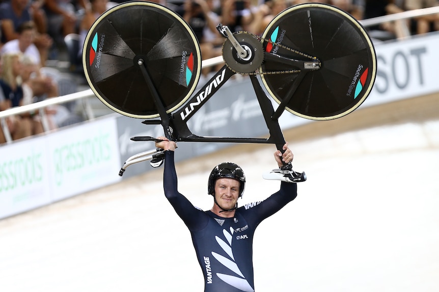 A man wearing black lycra and a black helmet holds a bike above his head