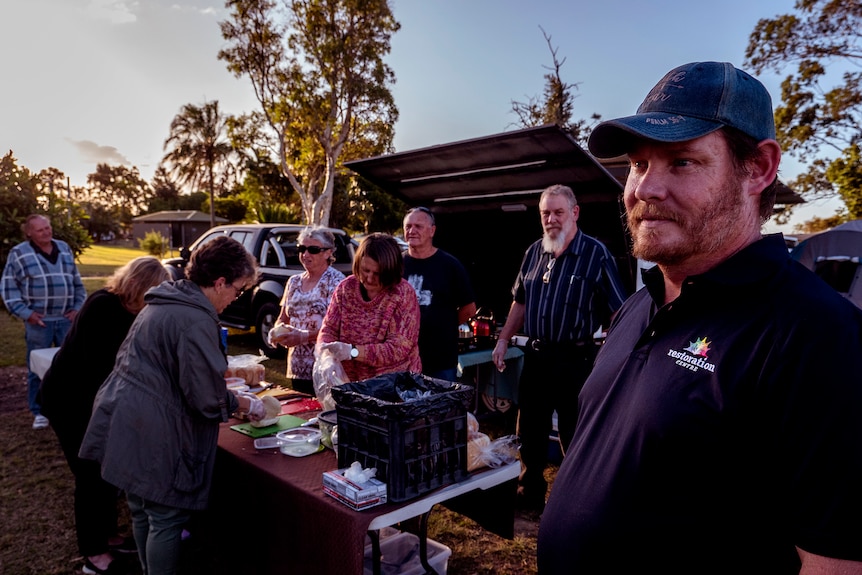 A group of people prepare food over a barbecue, while a bearded man with a cap is in the foreground.