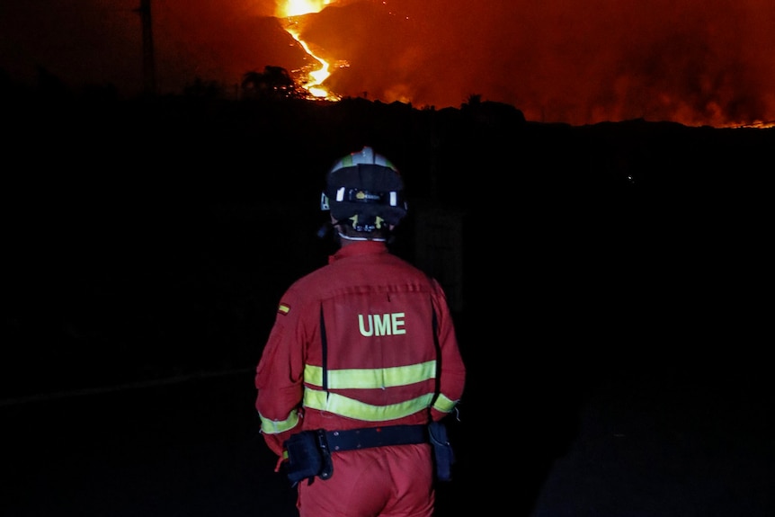A fireman ir orange looks at a burning volcano in the distance at night.