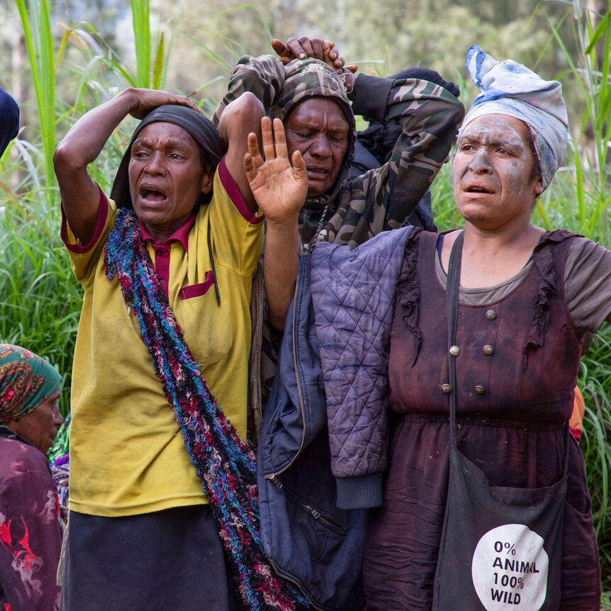 Four women stand with their hands up wailing in sadness with long green grass behind them