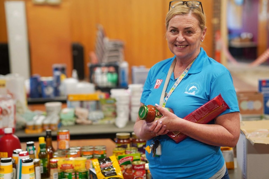 A woman holds a packet of pasta and a jar of pasta sauce standing in front of tables of groceries in a community hall.