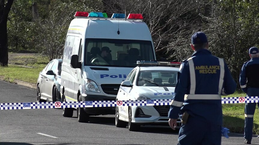 ambulance officers stand near police tape on a street in Nowra with police vehicles parked behind the tape