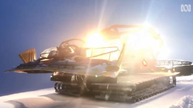 A snowcat with lights blazing on snow with a blue sky in the background.