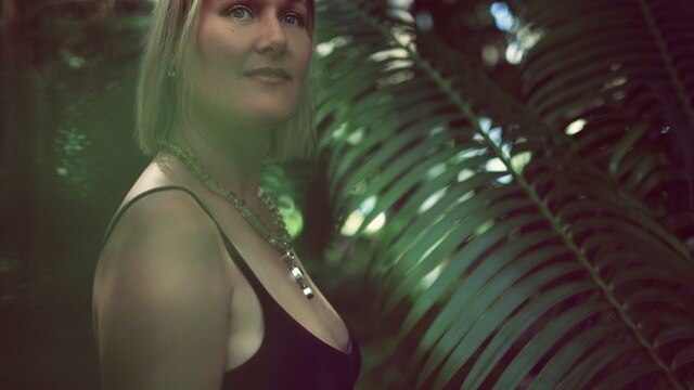 A light-haired woman  wearing a black singlet with ferns in background