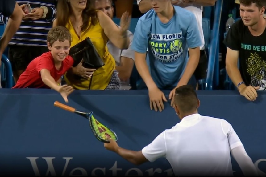 Kyrgios hands a clearly broken racquet to a fan, who is leaning over the fence and smiling.