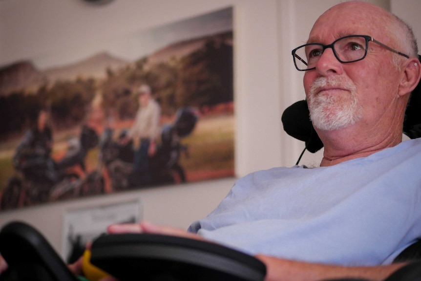 Tim Trembath sits in his wheelchair in his loungeroom in front of a portrait of him and his wife with their motorbikes
