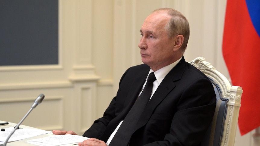 Russian President Vladimir Putin is pictured sitting at a desk, wearing a black suit and tie. Behind him is the Russian flag.