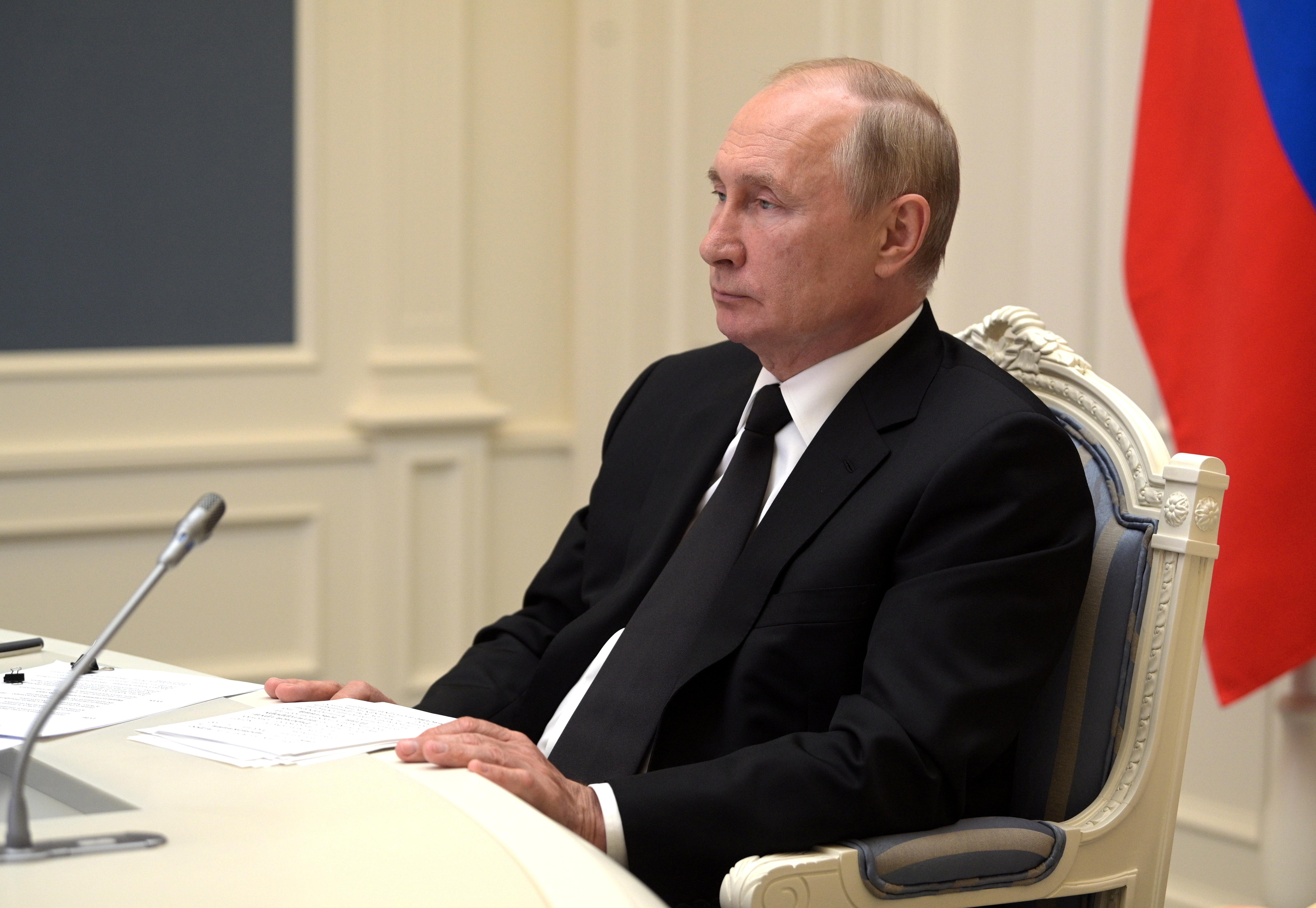Russian President Vladimir Putin is pictured sitting at a desk, wearing a black suit and tie. Behind him is the Russian flag.