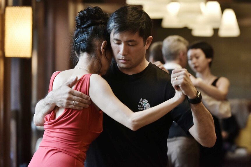 A young man with dark hair and a woman in a red dress intimately dance the tango together.
