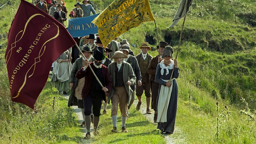 Colour still of a group of marchers with banners and dressed in Sunday best walking through field in 2018 film Peterloo.