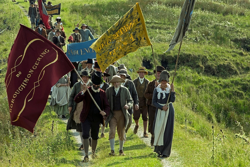 Colour still of a group of marchers with banners and dressed in Sunday best walking through field in 2018 film Peterloo.