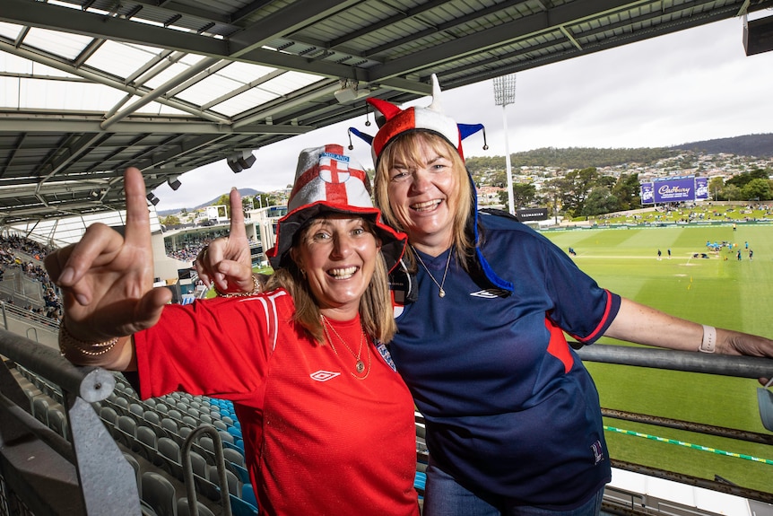Two women smile in the stands in front of a cricket oval.