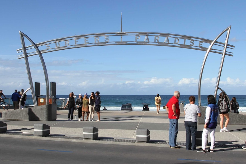 People standing under metal sign saying "Surfers Paradise" in front of a beach.