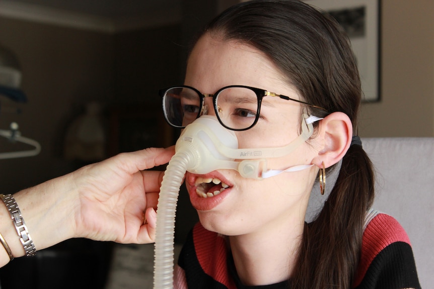 A close-up shot of Amy Evans on a ventilator with someone else's hands adjusting it on her face.