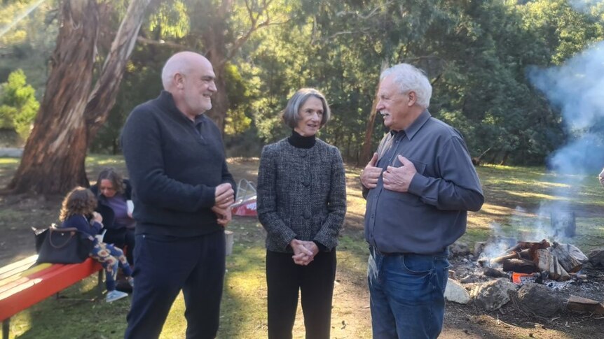 A woman flanked by two older men talking in a clearing with the sun dappling through trees, campfire in background
