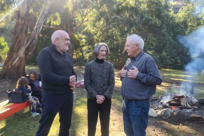 A woman flanked by two older men talking in a clearing with the sun dappling through trees, campfire in background