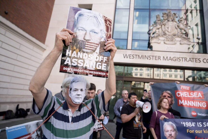 Someone wears a mask of Assange's face and holds a large poster of him outside court.