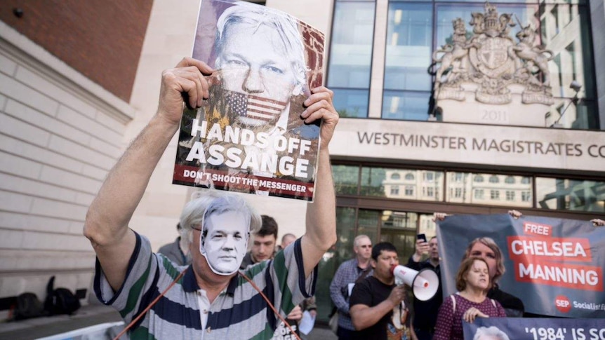 Someone wears a mask of Assange's face and holds a large poster of him outside court.