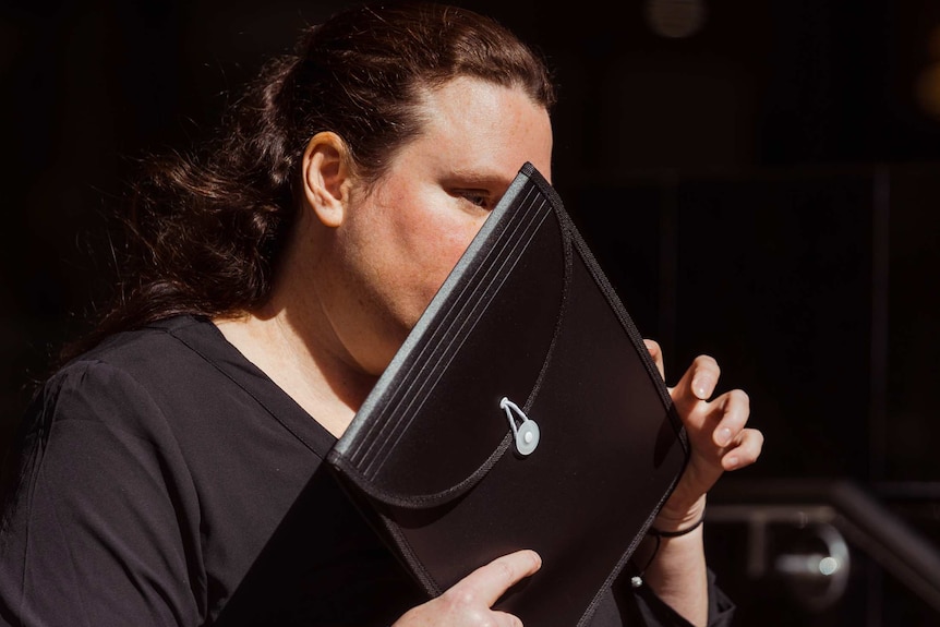 A woman in black holds a black folder over her face.