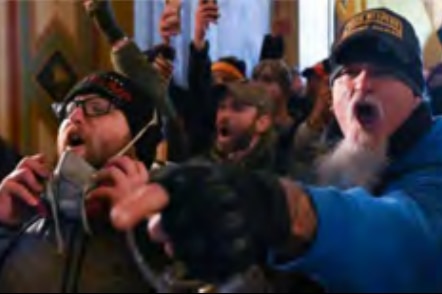 A man with a long white goatee and fingerless gloved yells in a hall full of rioters.