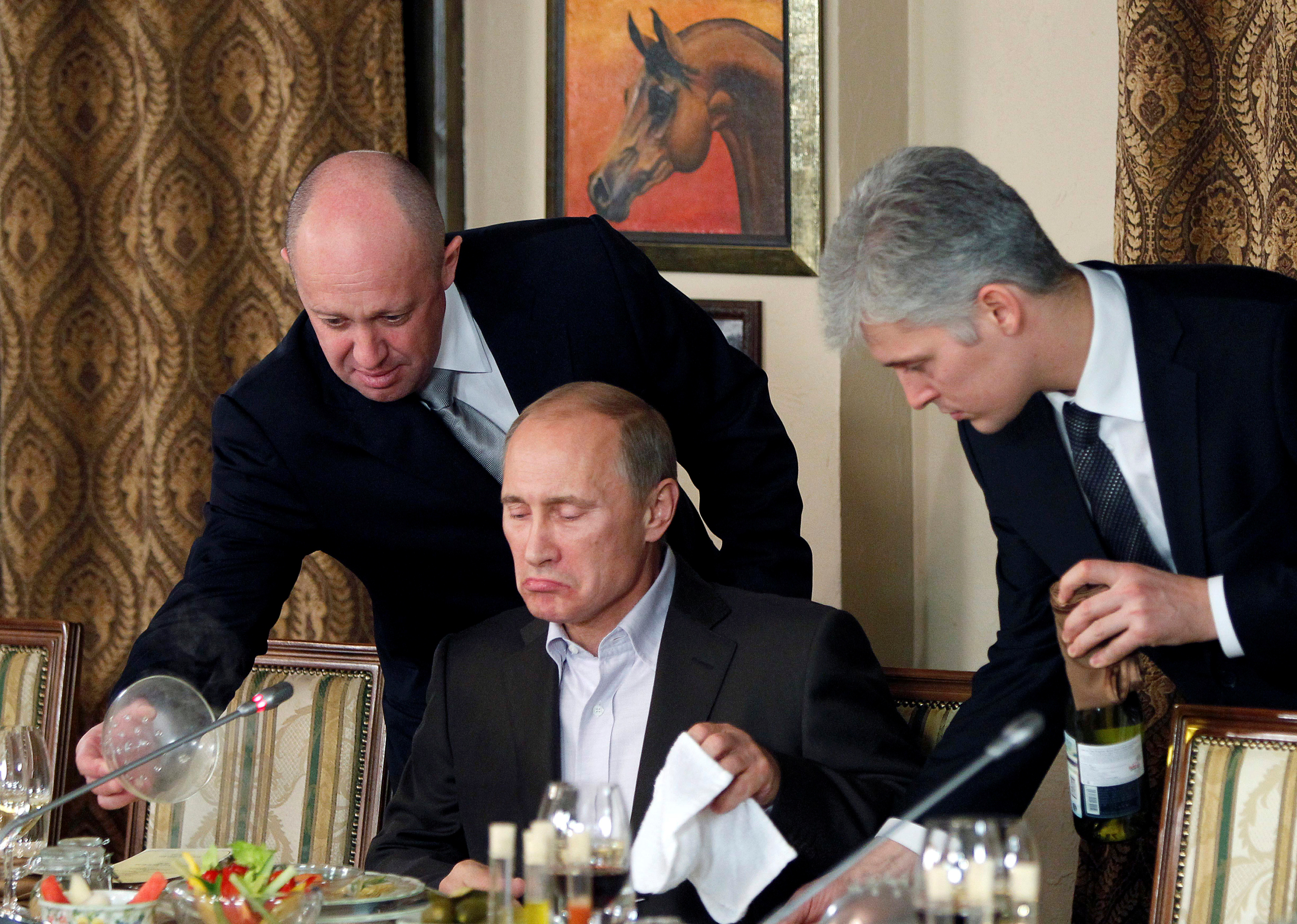 T两个男人站在俄罗斯总理弗拉基米尔·普京身边，因为他正准备开始用餐。” class=