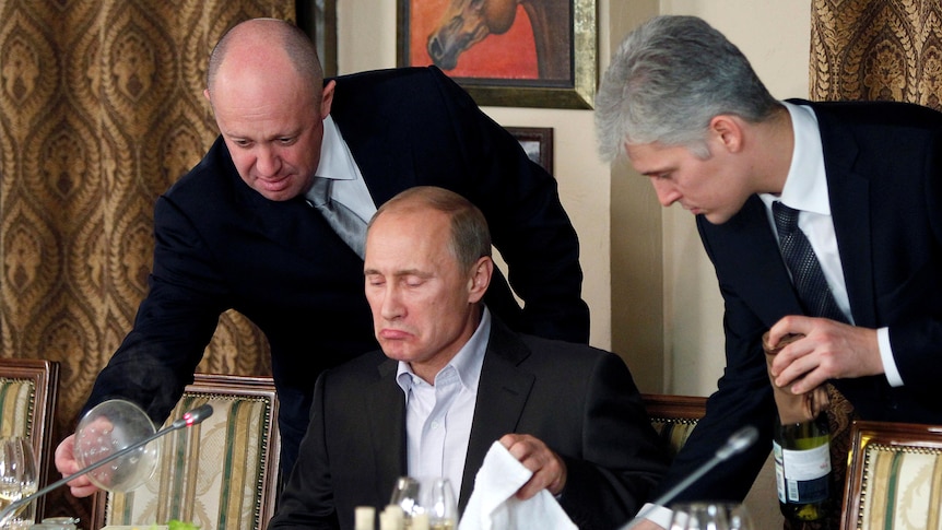 Two men stand over Russian Prime Minister Vladimir Putin as he is about to start a meal.