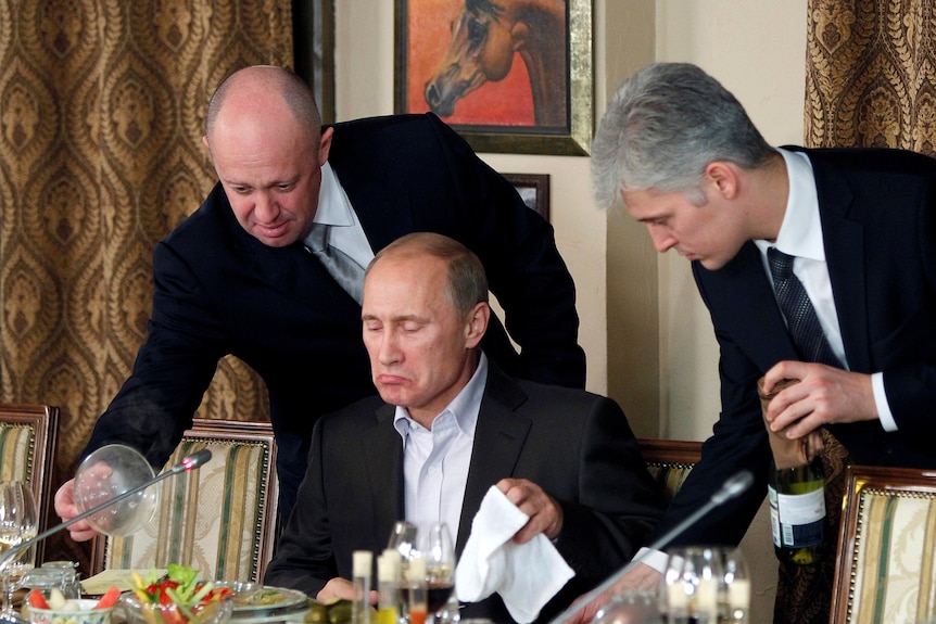 Doi bărbați stau în fața președintelui rus Vladimir Putin în timp ce acesta este pe cale să mănânce.