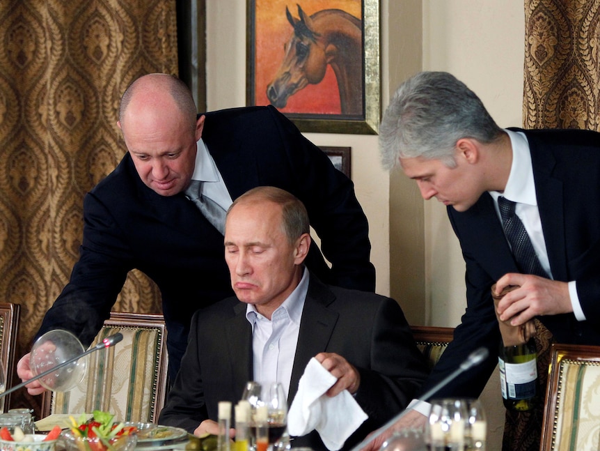 Two men stand over Russian Prime Minister Vladimir Putin as he is about to start a meal.