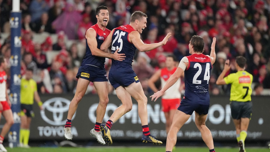 An AFL player jumps in the air in celebration as his teammates react after a goal.