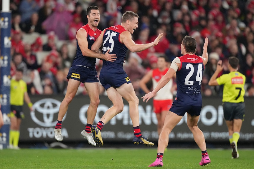 An AFL player jumps in the air in celebration as his teammates react after a goal.