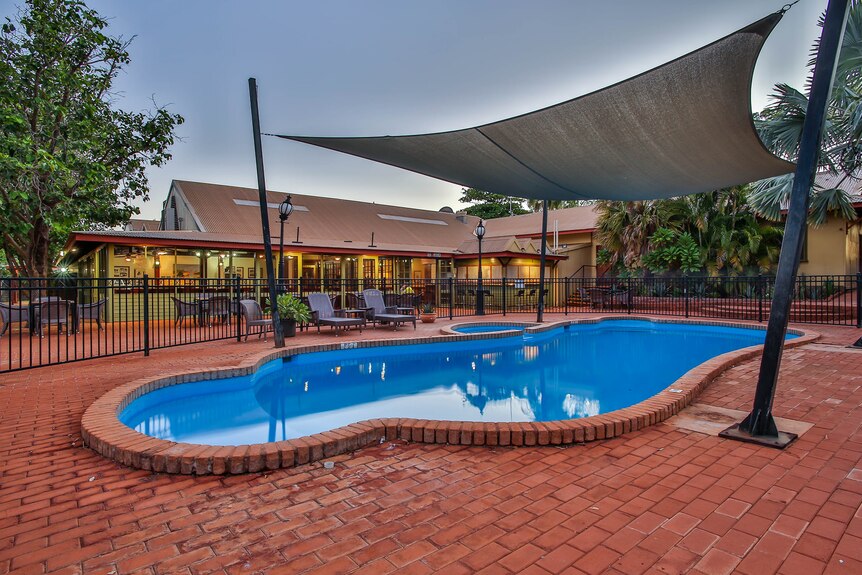 A hotel pool surrounded by red brick floor, a canvas shade and a fence. Another building can be seen in the distance.