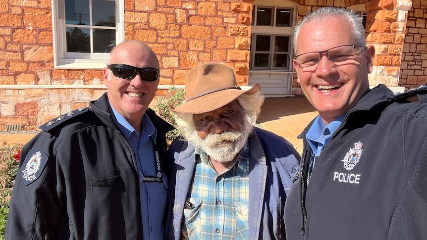 Selifie of two smiling bald, white men in police jackets with Indigenous elder, white beard, wears hat, outside brick building.