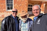 Selifie of two smiling bald, white men in police jackets with Indigenous elder, white beard, wears hat, outside brick building.