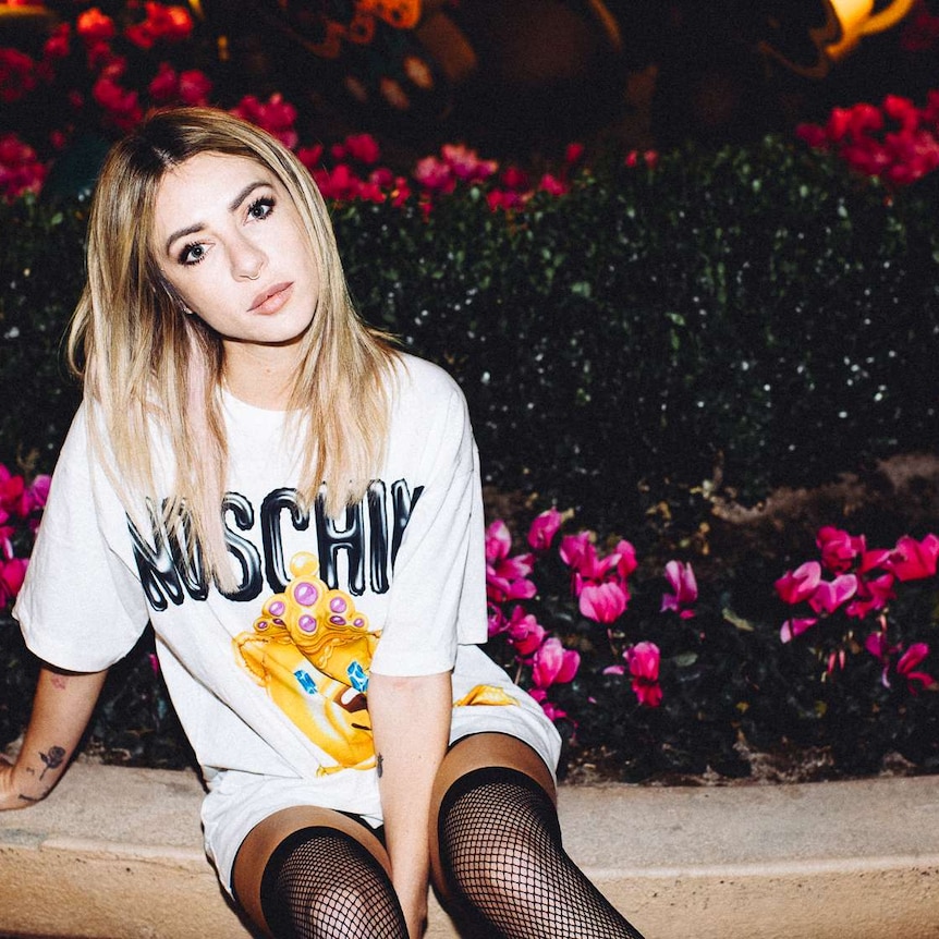 alison wonderland sitting on the edge of a flower garden at night, she's wearing a moschino tshirt and thigh high fishnets