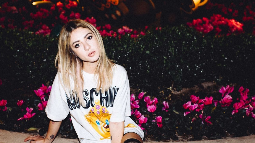alison wonderland sitting on the edge of a flower garden at night, she's wearing a moschino tshirt and thigh high fishnets