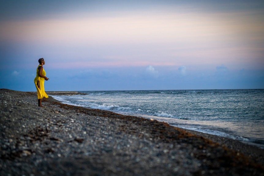 A woman in a bright yellow dress stands at the ocean's edge, taking in a beautiful sunset