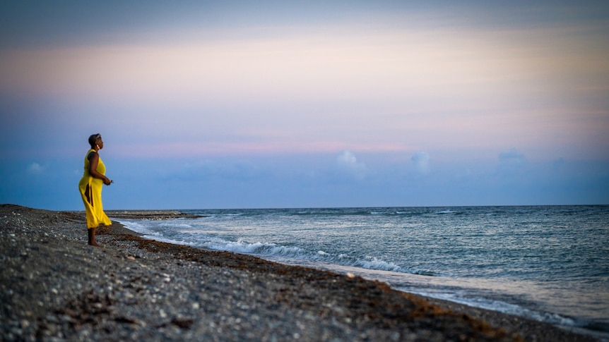 A woman in a bright yellow dress stands at the ocean's edge, taking in a beautiful sunset