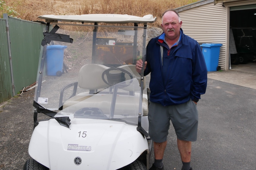 a man in a blue jacket stands next to a golf cart with a smashed windscreen 