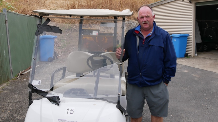 A moustachioed man in a dark jumper stands next to a smashed-up golf buggy.
