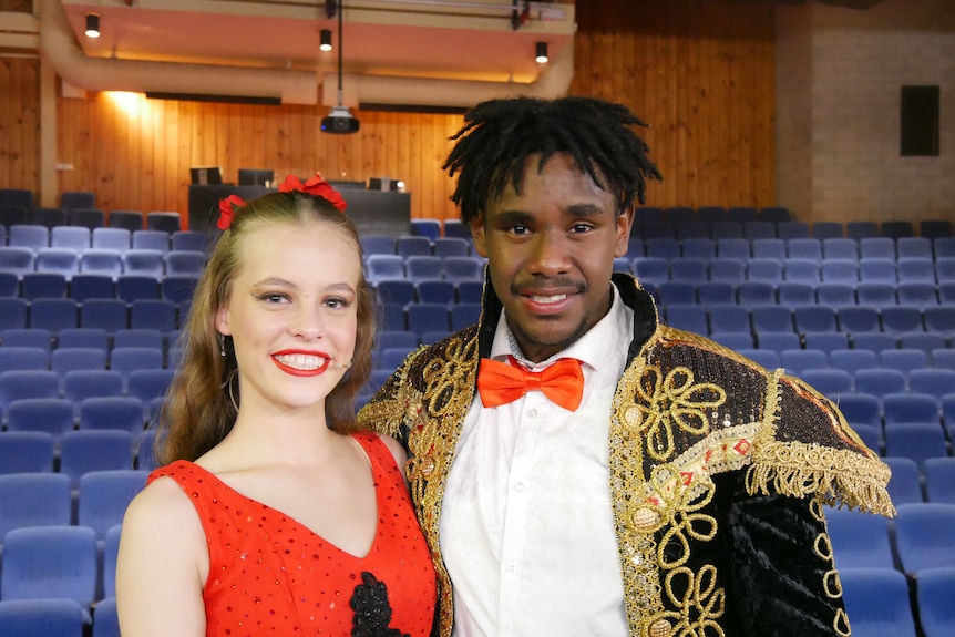 A young woman and man wearing stage costumes stand arm in arm and smile at the camera.