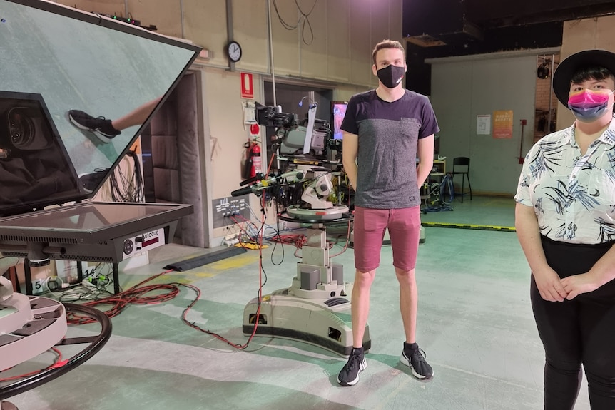 Two people wearing masks stand next to tv broadcasting equipment including a camera