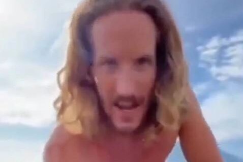 Blurred still taken from a video of a man with long curly hair, staring into camera, from shirtless waist up, under blue sky.