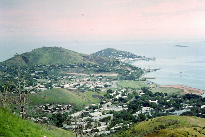 A view from a height of green mountains leading down to the sea, with clumps of buildings strewn across them.